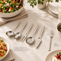 A 5-piece stainless steel serving utensil set arranged on a beige tablecloth with a salad bowl, plates, and dining props around it. The set includes a serving spoon, a serving fork, a decorative serving spoon, a cake server, and a dinner fork.