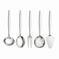 A 5-piece stainless steel serving utensil set arranged on a beige tablecloth with a salad bowl, plates, and dining props around it. The set includes a serving spoon, a serving fork, a decorative serving spoon, a cake server, and a dinner fork.
