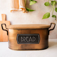 A rustic bronze-colored metal bread bin with a wooden lid and a black metal label that reads 'BREAD'. The bread bin is placed on a white surface with a textured beige wall in the background, decorated with a wooden cutting board, a beige cloth, and green plant leaves.