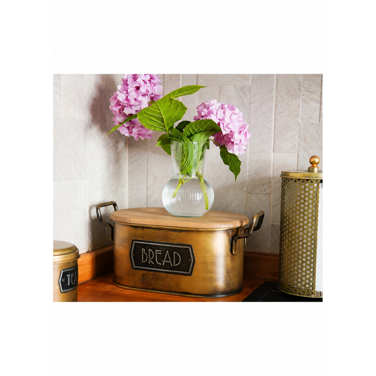 A rustic bronze-colored metal bread bin with a wooden lid and a black chalkboard label reading 'BREAD', styled with a glass vase of pink hydrangeas on a wooden countertop against a tiled wall.