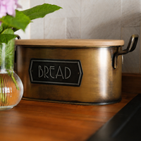 A rustic metal bread box with a light wood lid and a black label reading 'BREAD', placed on a wooden countertop with a tiled wall behind it and a vase of flowers at the side.