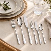 A 6-piece stainless steel flatware set arranged on a beige marble table runner, styled with matching plates, a glass, and a napkin.