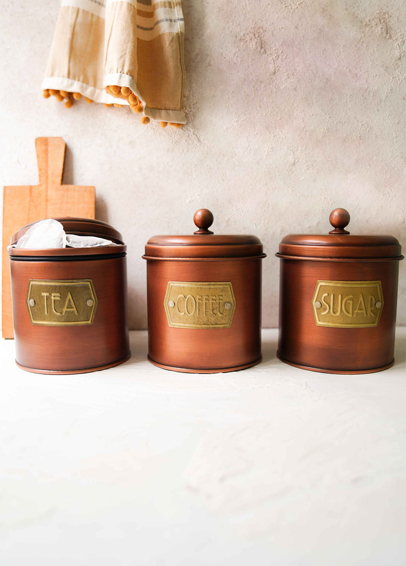 A set of three copper-colored galvanised steel canisters with labels Coffee, Sugar, and Tea on them, designed for storing coffee, tea, and sugar.