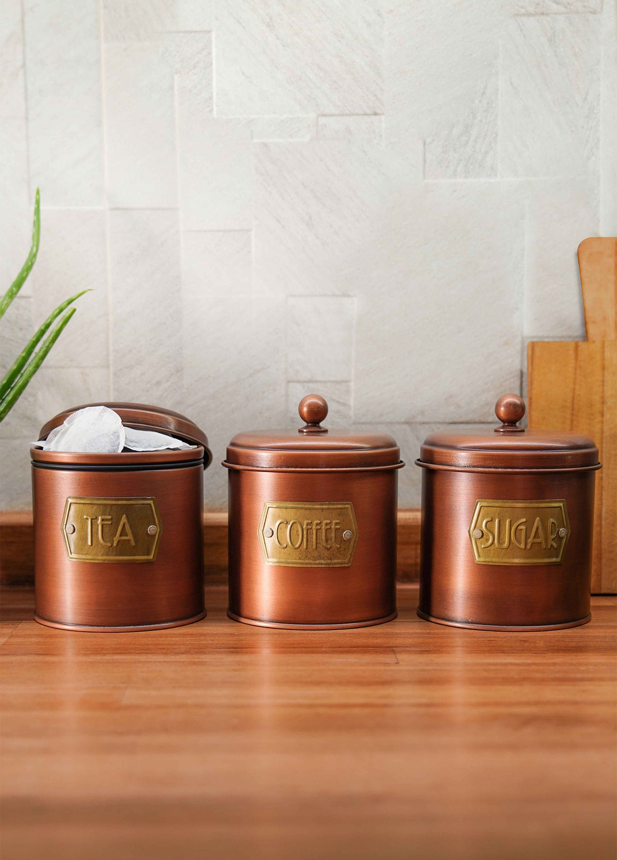 A set of three copper-colored galvanised steel canisters with labels Coffee, Sugar, and Tea on them, designed for storing coffee, tea, and sugar.